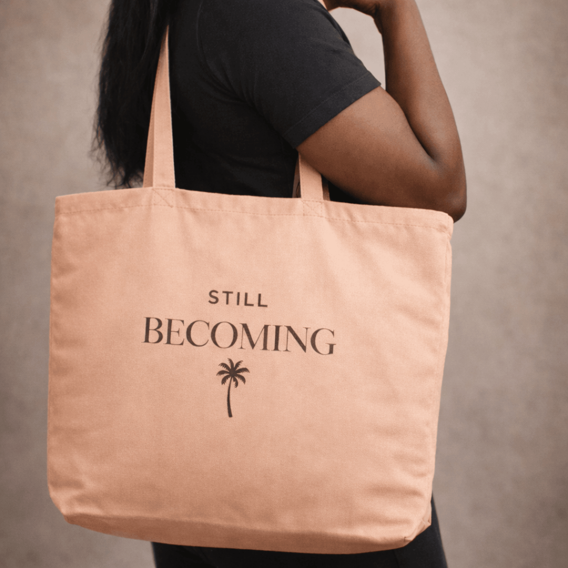 Person holding a tan tote bag with 'STILL BECOMING' text and palm tree design on a neutral background